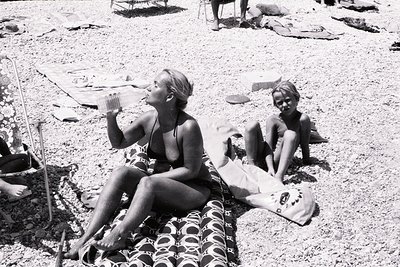Mid-20th century beach scene featuring two women lounging on patterned towels. The woman on left holds a drink, wearing a two...