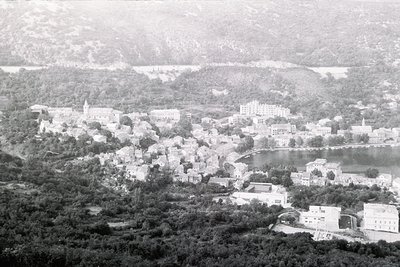 Aerial view of a coastal town nestled between hills and a lake, mid-20th century. Dense clusters of low-rise buildings with f...
