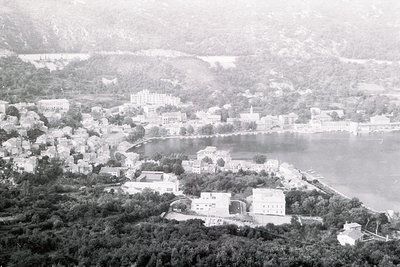 Aerial view of a lakeside town with mid-20th century architecture, likely . Dense residential buildings cluster along the wat...