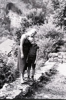 A mid-20th-century black-and-white photo of a woman and child posing on rocky terrain beside a historic alpine fortress. The ...