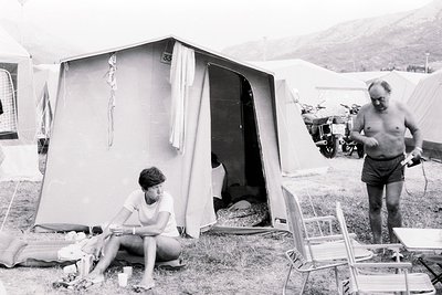 1970s-era camping scene with vintage canvas tent. Man in shorts reads newspaper near motorcycle; woman sits on ground near te...