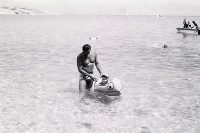 Mid-20th century seaside scene: man assists child in inflatable toy boat in shallow waters, with another boat and people in b...
