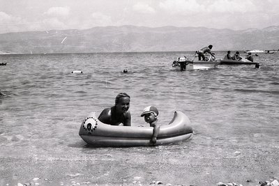 Vintage black-and-white seaside scene featuring two individuals in an inflatable raft near shallow waters. In background, two...