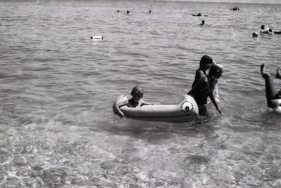 Black-and-white seaside scene: two adults and a child on an inner tube in shallow water, surrounded by swimmers. Mid-20th cen...