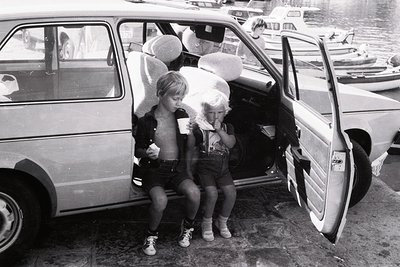 Two children in 1970s beachwear—shorts, tank tops, and sandals—sit on an open hatchback door, each holding a large ice cream ...