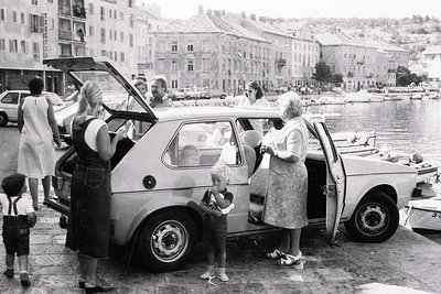A 1970s seaside scene: group of people loading luggage into a compact hatchback near a waterfront promenade. Mid-rise Soviet-...