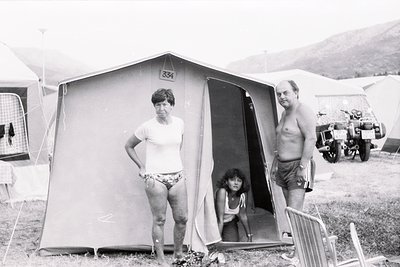 Family poses outside a numbered camping tent () in a grassy campsite, likely 1970s. Adults in swimwear, child seated in tent ...