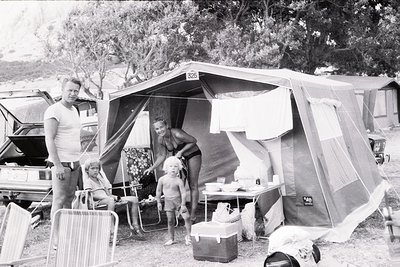 Family camping scene in a vintage tent () with classic folding chairs and a cooler, likely mid-20th century (). Man in tank t...