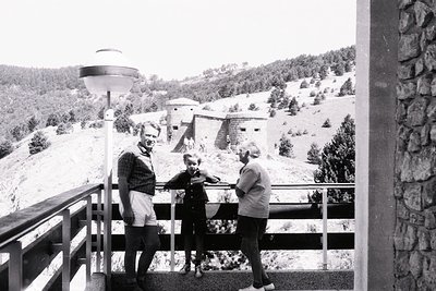 Three adults pose on a wooden balcony overlooking a fortified hillside complex, likely a Cold War-era bunker system. Mid-cent...