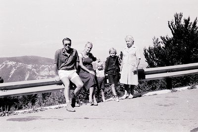 Family portrait on a mountain road, 1960s. Four individuals—man in plaid shirt/sunglasses, woman in sleeveless dress, child i...