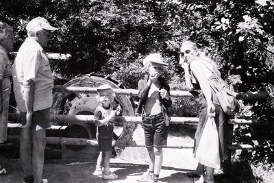 Mid-20th century outdoor scene featuring four adults and two children near a vintage amusement ride. Adults wear 1950s–60s fa...