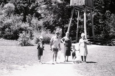 Mid-20th century family portrait in a park setting, likely 1950s–1960s. Five individuals pose near a wooden observation tower...