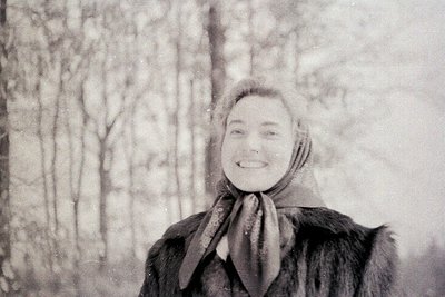 Vintage sepia-toned portrait of a woman in winter attire, standing in a snow-covered forest. She wears a fur-lined coat, scar...