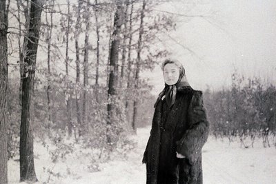 Black-and-white winter portrait of a woman in a dense forest, bundled in a long fur coat and headscarf. Snow blankets the gro...