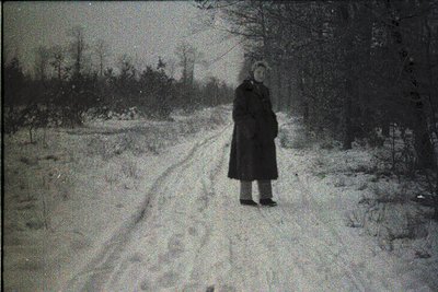 A lone figure in a long winter coat stands on a snow-covered path flanked by bare trees, evoking early 20th-century rural sol...