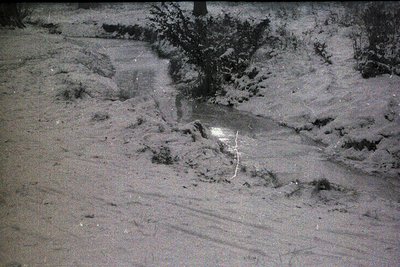 Vintage black-and-white photo of a snow-covered landscape with tire tracks and a faintly visible road. Snowbanks line a narro...
