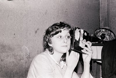 Vintage black-and-white photo of a person using a large, retro film camera, likely a Leica or similar. Indoor setting with te...