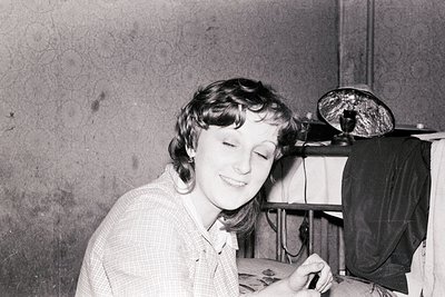 Mid-20th-century black-and-white photo of a woman in a checkered blouse, seated indoors with a vintage radio or speaker behin...
