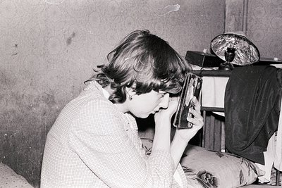 Vintage black-and-white photo of a person using a vintage rotary phone, seated indoors with a lamp and wooden furniture. Mid-...