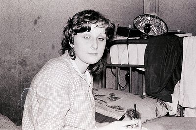 Portrait of a woman seated indoors, likely in a 1960s–1970s studio setting. She wears a checkered blouse and poses with a neu...