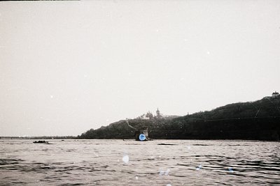 Black-and-white coastal scene featuring a lone figure in a small boat on calm waters, likely fishing or rowing. In the backgr...