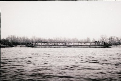 Black-and-white shot of a long, low-lying industrial dock with freight trains and warehouses along a waterfront. The scene su...