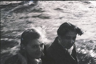 Black-and-white candid shot of two men aboard a boat, facing the churning sea. Both wear dark, textured jackets and short hai...