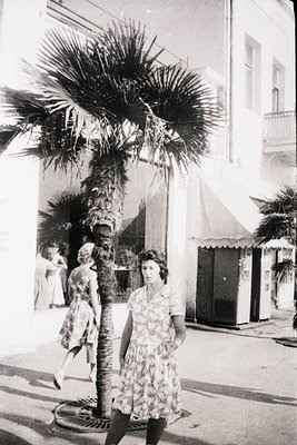 Mid-century seaside promenade with palm trees lining a wide sidewalk. A woman in a floral dress poses near a manhole cover, w...