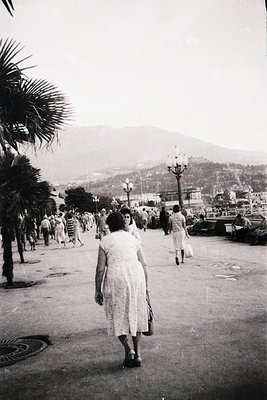 A mid-20th century seaside promenade bustling with summer crowds. A woman in a lace dress walks away from the camera, palm tr...