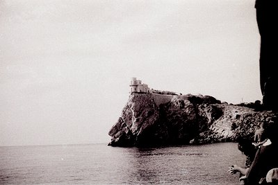 Black-and-white coastal scene featuring a lone, rectangular stone watchtower perched on jagged cliffs overlooking the sea. Mi...