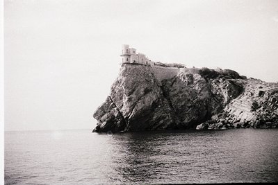Monolithic fortress perched on rugged cliff edge overlooking misty sea. Architectural ruins feature cylindrical towers and cr...