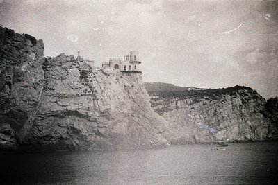 Black-and-white coastal scene featuring a cliffside fortress perched on jagged rock formations overlooking a calm sea. Archit...
