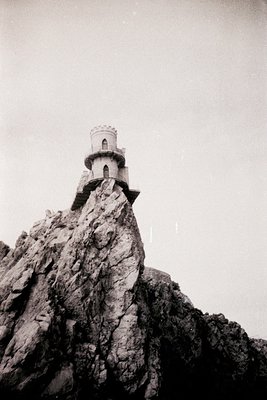Historic lighthouse perched atop rugged cliff, likely mid-20th century. White cylindrical tower with conical roof, set agains...