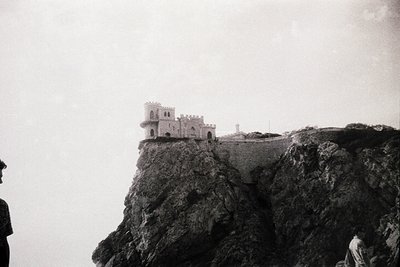 Historic fortress perched atop rugged cliffs, likely a coastal watchtower or medieval stronghold. Black-and-white photograph ...