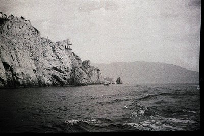 Black-and-white coastal scene featuring jagged cliffs rising from choppy waters. A small, elevated structure perched atop the...
