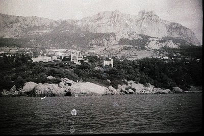 Mid-20th century coastal town nestled between rugged cliffs and alpine peaks, likely Mediterranean. Prominent seaside archite...
