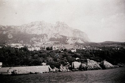 Vintage coastal town nestled between rugged cliffs and snow-capped mountains, likely Mediterranean. Prominent stone fortifica...