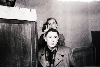 Vintage black-and-white portrait of two men in a dimly lit indoor setting, likely mid-20th century. Foreground subject wears ...