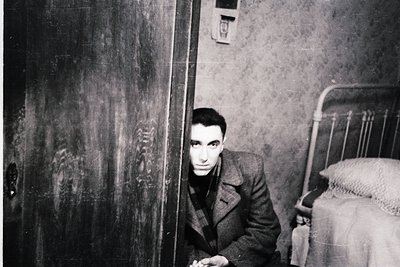 A man in a dark coat sits on a bed in a dimly lit, minimally furnished room, peering through a slightly ajar door. The interi...