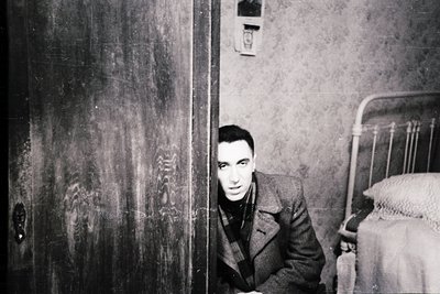 Mid-century man in a dark coat peeks from a dimly lit doorway, framed by worn plaster walls and a partially visible bed. The ...