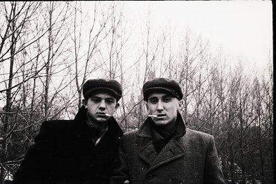 Two men in mid-20th-century winter attire pose outdoors in a leafless forest. Both wear flat caps, overcoats, and scarves; on...