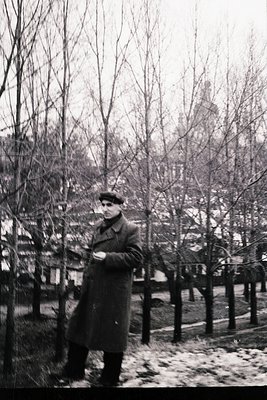 Mid-20th century man in winter coat and cap stands on snow-dusted path flanked by bare trees. Urban park or residential area ...
