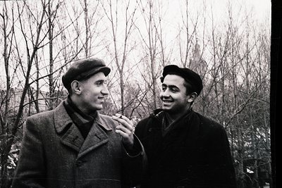 Two men in mid-20th century attire pose outdoors in a leafless park, likely Eastern Europe. The man on left wears a flat cap ...