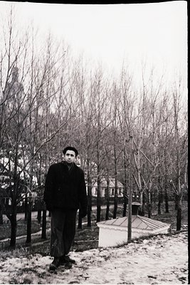 Black-and-white portrait of a man in winter attire—dark coat, scarf, and cap—standing on a snow-dusted park path. Leafless tr...