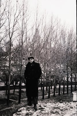Mid-century man in a winter coat and beret stands on a lightly snow-covered path, flanked by bare trees and a wooden fence. U...