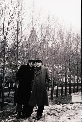 Two men in winter coats and flat caps pose outdoors in a park-like setting, likely late 1940s–1950s. Snow patches on ground s...