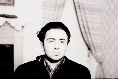 Portrait of a young man in mid-20th century formal wear—dark blazer, checkered shirt, and tie—posed indoors against a plain b...