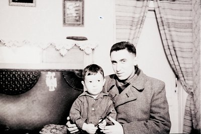 Vintage black-and-white portrait of a man and young boy in a modest indoor setting, likely mid-20th century. The man wears a ...