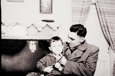 A man in a tweed overcoat and a young boy in a sweater pose indoors, likely mid-20th century. The man holds a toy train, whil...