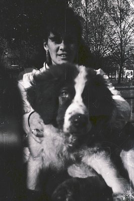 Vintage black-and-white portrait of a woman holding a large, shaggy dog in an urban park. Fur coat and dog’s coat suggest mid...